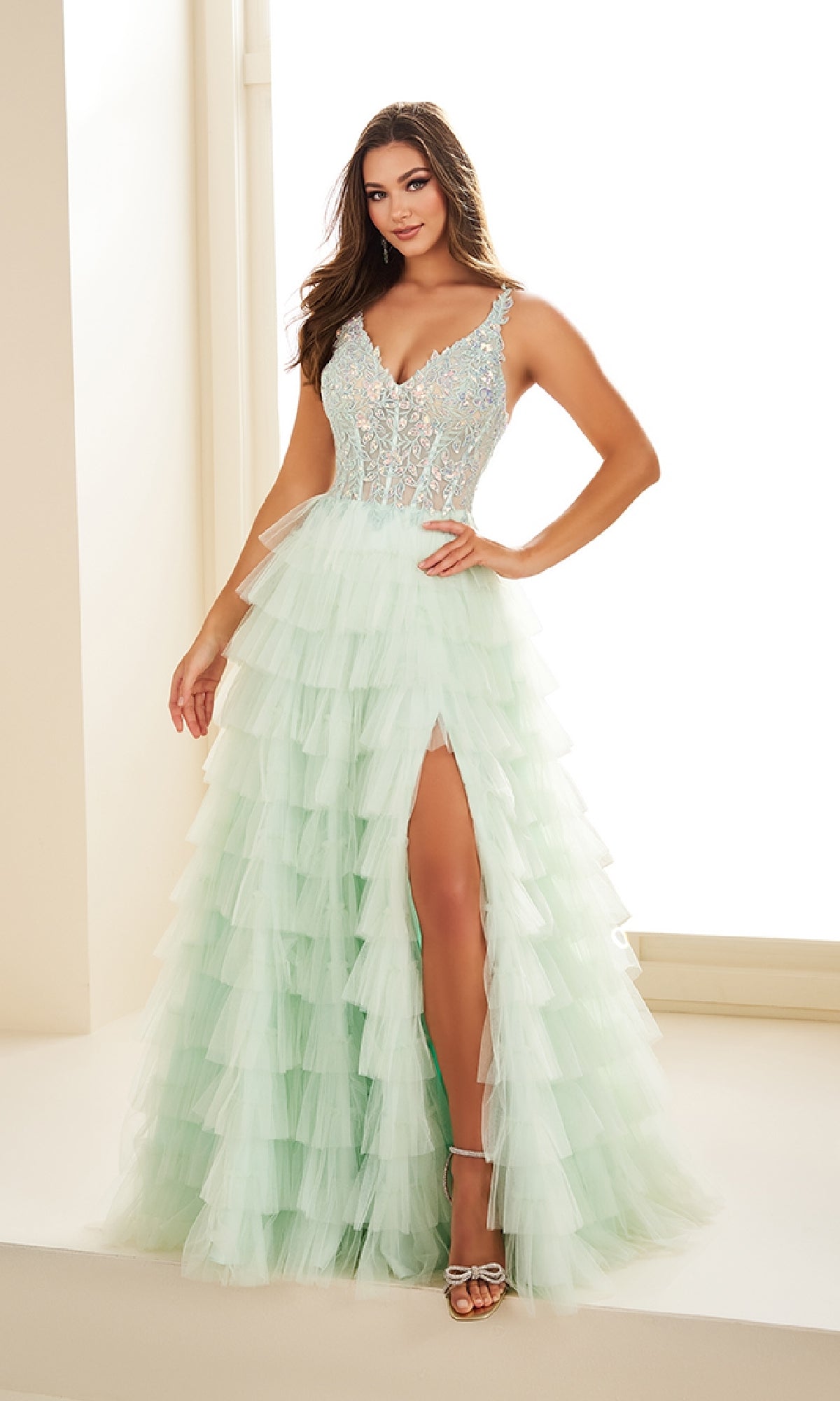 A woman wearing the Tiered Ellie Wilde EW35059 Designer Ball Gown with a lace bodice, thin straps, ruffled tiered skirt, and high leg slit stands indoors, smiling at the camera.